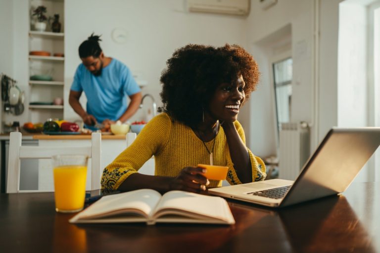 A multicultural woman is checking her bank account from home while her husband is cooking dinner.